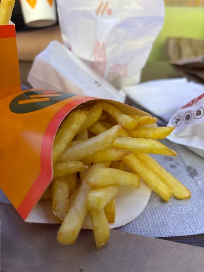 A close-up, eye-level shot shows a partially filled orange and red paper container of french fries. The fries are golden yellow and appear to be salted. The container is tilted towards the viewer, with the fries spilling out onto a gray, textured paper napkin. In the blurred background, there are white paper fast-food bags with orange and red lettering. The overall setting suggests a casual, outdoor dining environment, possibly at a fast-food restaurant. There is no indication of Shendzhii, Russia.