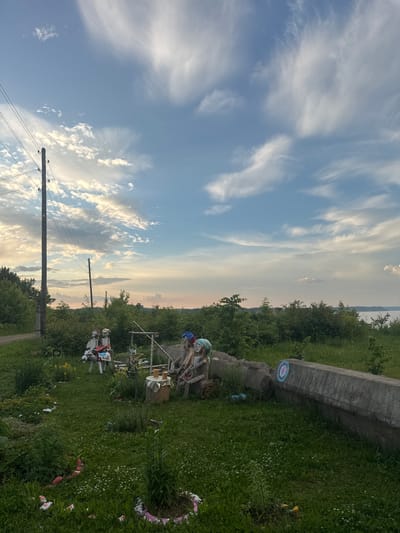 The image depicts an outdoor scene with a backdrop of a cloudy sky and a body of water in the distance, likely overlooking the Volga River. In the foreground, an assortment of discarded objects and handmade figures are scattered across a grassy area. A tall utility pole with wires stands to the left, and a dirt road is visible adjacent to it.

Dominating the mid-ground are several makeshift figures resembling scarecrows or folk art creations. One group of figures appears to be seated around a small table with what looks like mugs. Another set of figures is arranged on a wooden structure. To the right, a long concrete structure is partially visible, with a painted circular target on its surface. The overall impression is one of a rural or semi-abandoned outdoor space with a touch of whimsical, handmade artistic expression. The location is identified as Chastyye, Russia.