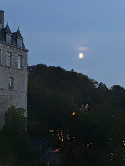 The image shows a nighttime scene with a partial moon visible in the dusky blue sky. On the left, a stone building with a mansard roof and dormer windows is prominent. To the right of the building, a densely wooded hillside rises. A small, ornate building with a red roof and a pointed tower is nestled among the trees on the hillside. A streetlamp illuminates a patch of the trees. The overall impression is of a tranquil evening setting in or near the historic town of Durbuy, Belgium.