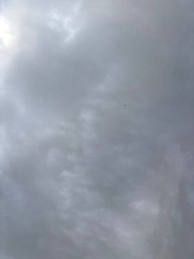 A photograph depicts a cloudy sky. There are a few very small, dark specks, potentially birds or insects, visible in the upper left and central portions of the frame. The overall tone of the image is grey, suggesting overcast conditions. The location is specified as Mārupe, Latvia.