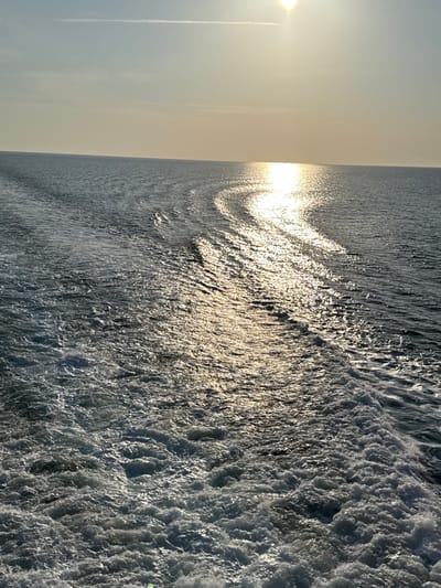 The image depicts an open maritime scene, geographically located in the area of Palaiopoli, Greece. A clear, light yellowish-blue sky occupies the upper portion, featuring a faint, linear contrail in the upper-left quadrant. The sun, appearing as an intense bright light source, is situated in the upper right, casting a broad, golden-white shimmering reflection that extends in an undulating path across the water towards the viewer. The majority of the lower frame is dominated by a substantial, turbulent white and foamy wake, indicative of a moving vessel, churning the dark blue-grey sea. No landmasses, artificial structures, or individuals are visible.