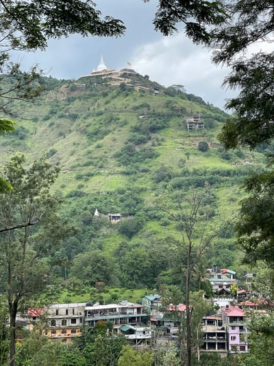 The image shows a hillside in Kumbalwela, Sri Lanka. The hillside is primarily green with vegetation. A white stupa (Buddhist monument) is visible at the top of the hill. Scattered buildings are seen along the hillside and at the base of the hill. Trees frame the top and bottom of the image. The sky is overcast.