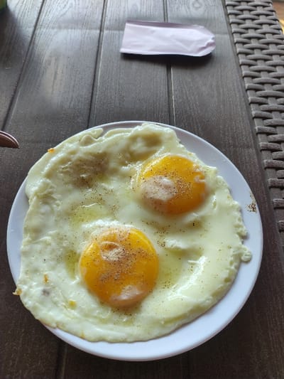 The image shows two fried eggs on a white plate, with black pepper sprinkled on top. The eggs are on a wooden table. There is a pink rectangular object to the left of the plate. The location of the image is in Gambiran, Indonesia, at coordinates -8.434 latitude and 114.18 longitude.