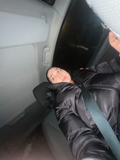 A person in a black puffer jacket is seated in a car. The interior is gray, and the car appears to be moving at night. A seatbelt is fastened across the jacket. The person is looking towards the camera with a neutral expression. The perspective is slightly angled. A portion of the car's ceiling and the outside night view are visible. The sun visor on the driver's side is down.