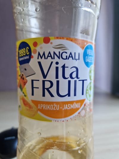 The media file shows a close-up, horizontal shot of a clear plastic bottle containing a light-colored liquid, likely a beverage. The bottle is partially filled, with condensation visible on its surface, suggesting it might be cold. The label on the bottle is the primary focus.
The label is predominantly white with colorful graphics and text. Prominently displayed is the brand name "MANGAĻI" and the product name "Vita FRUIT" in large, dark blue sans-serif letters. To the left of "MANGAĻI" are two circular badges, one blue with white text reading "VIEŅU GAZE" and another green badge with illegible text. To the right of "Vita FRUIT", also on an orange background, is text that reads "APRIKOŽU - JASMĪNU", indicating the flavor. Below the "Vita FRUIT" text, there are illustrations of what appear to be fruit slices, possibly apricot and some green leaves, along with a grey, stylized representation of a laptop or electronic device.
In the lower left corner of the label, a dark blue circular badge contains the text "7,999 €" in yellow, with smaller white text below it reading "Galvenā balva!".
The background is blurred and out of focus, appearing to be an indoor setting, possibly a table or counter. The lighting suggests it is daytime. There are no people visible in the image. The overall impression is a product shot focusing on the branding and flavor information of the beverage.