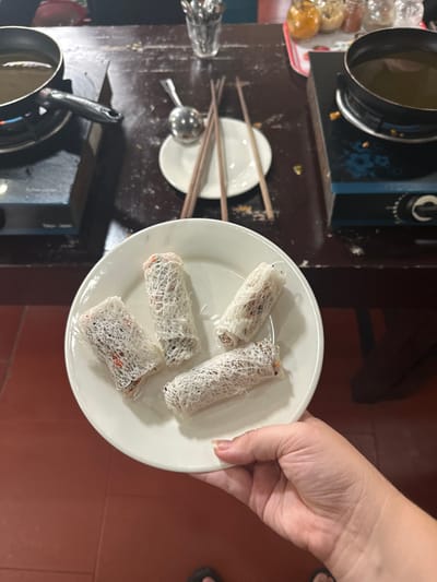 A white plate holds four spring rolls covered in a thin, web-like rice paper. The rolls are arranged towards the top right of the plate. A hand holds the plate. In the background, a dark wooden table displays various cooking items, including a hot pot, a small pot on a burner, a white plate with chopsticks, a spoon, and a glass. The floor is red tile.