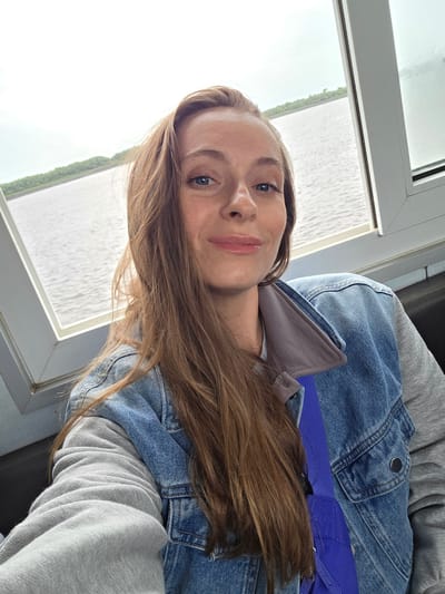 This close-up selfie, originally rotated 90 degrees counter-clockwise, features a young woman with long, straight, light brown hair and blue eyes. She is positioned indoors, looking directly at the camera with a subtle, pleasant smile. Her attire consists of a light blue denim jacket worn over a grey hooded sweatshirt or top. A bright blue strap, possibly from a bag or a seatbelt, is visible across her chest.
Through a large, white-framed window to her left, a serene body of water is visible, likely a river or lake, reflecting the bright, overcast sky. A strip of green trees or land can be seen in the distance across the water. The weather appears to be cloudy, providing soft, even lighting. The setting suggests she is seated near a window, possibly on a boat, train, or in a building overlooking the water, in Leninskoe, Russia. The overall mood conveyed is calm and relaxed. There is no visible text in the image.