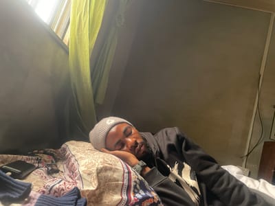 The image shows a person sleeping in a dimly lit room. The subject, a young Black man wearing a grey beanie and a black hoodie with a graphic design, is lying on a bed with a patterned duvet. His head is resting on a pillow, and his eyes are closed. A watch is visible on his left wrist. To the left of the bed, a black object, possibly a phone or wallet, rests on the duvet. Dark blue and purple knitwear items are also visible on the bed.

The room appears to be a bedroom, with a window covered by a light green curtain on the left side of the frame. The wall behind the subject is a neutral, textured grey. The lighting suggests it could be daytime, with natural light filtering in through the window, or it could be artificial lighting that is dim. The overall mood is one of rest and quiet. There is no visible text in the image. The location context is Aiyegunle, Nigeria.