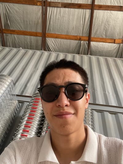 A close-up, slightly upward-angled selfie shows a young East Asian person wearing dark, round-framed sunglasses and a light-colored, textured polo shirt. The person's short, dark hair is visible above the sunglasses. The background consists of corrugated metal walls and what appears to be insulated sheeting on a ceiling, with wooden beams supporting the structure. To the left and behind the person, stacks of cylindrical metal objects, possibly ductwork, with visible red plastic separators, are arranged vertically. The overall environment suggests an indoor industrial or storage space, likely in a workshop or warehouse setting, possibly located in the village of Kenen Azirbayev, Kazakhstan.
