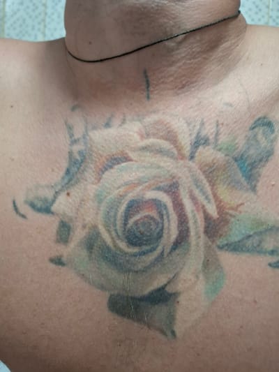 The image shows a close-up of a person's chest and neck area, featuring a tattoo of a rose. The tattoo is in muted colors, primarily pale pink, white, and blue-green, with darker shading. There is a thin black string or cord visible around the person's neck. The skin appears to have some texture and small blemishes. The background is out of focus, but suggests a tiled surface, possibly a bathroom wall. The location within Novlyanka, Russia, is not discernible from the visual information provided in the image.