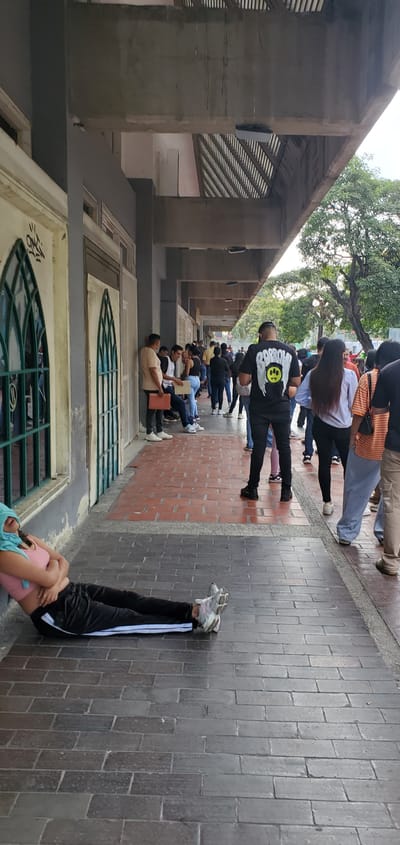 A street scene in Barquisimeto, Venezuela, shows a queue of people lined up against a building. The building has a series of arched windows with ornate metal grilles and a concrete overhang supported by columns. The people are standing or sitting along the brick-paved sidewalk, waiting in line. In the foreground, a person is lying on their back with their legs up against the wall. The street is shaded by trees and the building's overhang.