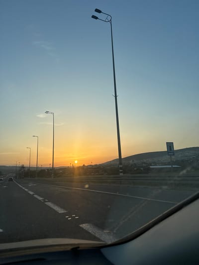 The image depicts an outdoor scene from the interior perspective of a vehicle on a multi-lane highway within the Merom HaGalil Regional Council, Israel, during either sunrise or sunset. The sky transitions from light blue at the top to orange and yellow at the horizon, with a distinct, bright sun visible on the left, slightly above the distant landscape.
The immediate foreground reveals the dark dashboard or cowl of the vehicle. The asphalt road surface occupies the lower half of the frame, marked with white dashed lane lines. A series of streetlights with single luminaires extends into the distance along the left side of the highway, with other vehicles faintly discernible. A prominent, tall streetlight featuring dual luminaires stands in the right mid-ground. Further along the right side of the road, a dark guardrail separates the highway from the adjacent terrain, where a blue roadside sign displaying a white gas pump icon and a right-pointing arrow is affixed to a pole.
The background consists of hills covered with vegetation, upon which scattered buildings and structures are visible, indicating a settlement or town.