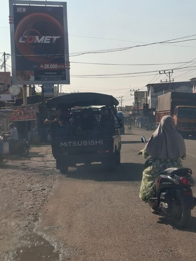The image captures a street scene in Blangmetimu, Indonesia. In the foreground, a woman wearing a light grey hijab and a green floral patterned dress is riding a black scooter. Behind her, a dark-colored Mitsubishi pickup truck is visible, with several people sitting in the open bed of the truck. The truck has a black tarp covering the top.

To the left of the truck, a man in a white shirt is riding a motorcycle. Further left, a stall with signage that appears to read "KHAS ACEH" is present, along with other structures that suggest a commercial area. Overhead, utility wires are strung across the sky, supported by poles. A large billboard advertising "COMET" with a price of "Rp. 26.000 (ISI 20)" and a "PERINGATAN" (warning) is prominently displayed on the left side of the image.

In the background, more buildings and utility poles line the street, and a large orange truck is parked on the right side of the road. The road surface appears to be asphalt and is partially wet, with puddles indicating recent rainfall or a water source. The overall lighting suggests it is daytime, with clear skies.
