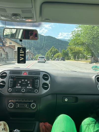 The interior of a car is visible, with the dashboard and infotainment system taking up the majority of the frame. Through the windshield, a view of a street in Kresna, Bulgaria, is visible. The road is straight, with buildings on either side and mountains in the background. Several cars are seen on the road. A VIP car freshener is hanging from the rear view mirror.