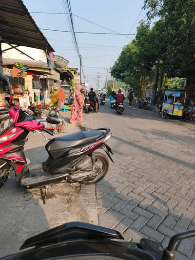 The image depicts a street scene in Sidorejo, Indonesia, likely during daylight hours given the shadows cast by the buildings and trees. The foreground is dominated by the front of a parked pink and black scooter. Beyond the scooter, the street is paved with interlocking bricks and shows signs of wetness.
On the left side of the street, a row of small shops or stalls lines the sidewalk. These structures have corrugated metal roofs and display various goods, including snacks and drinks on shelves. A motorcycle fuel pump is visible, suggesting one of the businesses might be a small convenience store or a gas station. Several people are present around these shops, some appearing to be customers or vendors.
The street itself is active with traffic. Several motorbikes are moving along the road, some carrying single riders and others with passengers. The people on the motorbikes are dressed casually, with some wearing headscarves.
Further down the street on the right, there is a food cart with a blue awning. People are gathered around this cart, and a man is seated nearby. A large tree with dense green foliage provides shade along the right side of the street. Overhead, a network of power lines crisscrosses the sky. The overall atmosphere suggests a typical day in a bustling urban or suburban area of Indonesia.