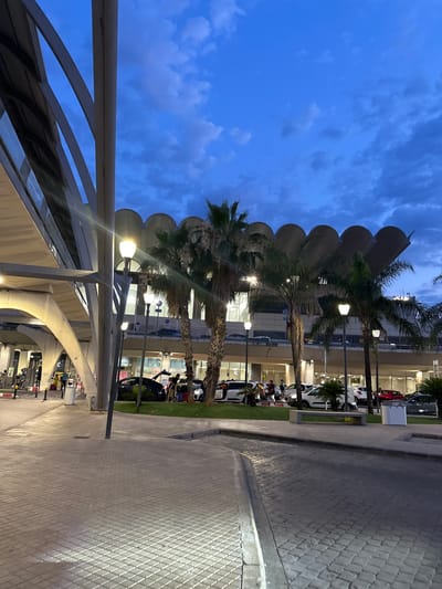 The image shows a view of the airport terminal in Manises, Spain at dusk. The foreground is a paved walkway and road, with some parked cars and people in the mid-ground. Several palm trees are scattered throughout the scene. The airport building itself is visible in the background, characterized by a distinctive roofline with rounded arches. The sky above is a deep blue, with scattered clouds, indicating the transition from day to night. Several streetlights are illuminated, casting light on the walkway. There are no discernible interactions between the people in the image.