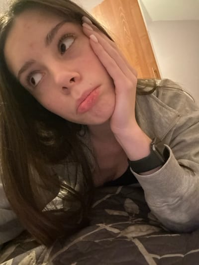 A woman lies on patterned bedding, angled to the left. She wears a grey zip-up jacket and an Apple Watch. Her face is partially obscured by her hand, which presses against her cheek. She is making a silly face, sticking out her tongue and crossing her eyes. Her dark brown hair is long and partially covers her shoulder. The background includes a glimpse of a light-colored wall and wooden surface.