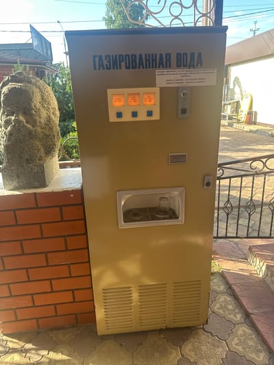 This is a medium-shot, eye-level, outdoor photograph of a vintage Soviet-era carbonated water dispenser in Kablukovo, Russia. The dispenser is a tall, beige, metal cabinet with Cyrillic text across the top that reads "ГАЗИРОВАННАЯ ВОДА" (Carbonated Water). Below the text, a smaller white label indicates that the machine accepts USSR coins and specifies prices for plain and flavored water. To the left of the label are two illuminated orange buttons, each labeled with a coin denomination and description: "БЕЗ СИРОПА МОНЕТА 1 КОПЕЙКА" (Without Syrup, 1 Kopeck Coin) and "С СИРОПОМ АПЕЛЬСИНОВЫЙ МОНЕТА 3 КОПЕЙКИ" (With Syrup, Orange, 3 Kopeck Coin). There is a coin slot in the center of the machine, and a glass is present in the dispenser area.
To the left of the dispenser, a rough, stone bust of a person's head and shoulders stands on a brick wall. In the background, a metal fence with ornate scrollwork is visible, with patterned paving stones on the ground. The scene appears to be in daylight, with shadows cast by the sun. The overall impression is one of a public space, likely an outdoor waiting area or a pathway.