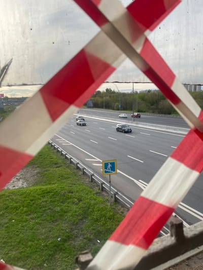 The image depicts a view of a highway in Вёшки, Russia, as seen through a barrier marked with red and white stripes. Several vehicles are visible on the highway. A pedestrian crossing sign is positioned alongside the road. Green grass covers the ground next to the highway.