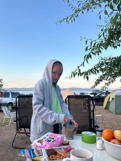 A young girl in a grey hooded sweatshirt and a light green shirt is standing at a table, stirring a drink in a clear glass mug with a blue utensil. She is looking at the camera with a neutral expression. On the table, there is a bowl of fruit (apples and an orange), a container of salt, a green container, a slice of bread, a package of pink cookies, and a package of pretzels. In the background, there are two chairs, tents, cars, and a body of water with hills in the distance. The scene appears to be a campsite. The location is identified as Kolodezny, Russia.