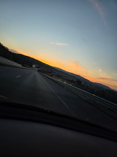 The image depicts a view from a vehicle driving on a highway at sunset. The sky is a gradient of light blue to orange, with a few wispy clouds. Rolling hills covered in trees line the side of the highway, with a small village visible in the distance. The highway itself has white lane markings and a metal guardrail. In the foreground, the dark interior of the vehicle is visible. The scene appears to be in or near Moshtanets, Bulgaria, judging by the landscape and road infrastructure.