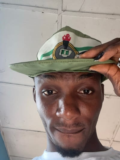 A low-angle, close-up selfie shows a young Black man wearing a green National Youth Service Corps (NYSC) cap. The cap has a white front panel with a circular logo featuring a torch, a map of Nigeria, and the text "NATIONAL YOUTH SERVICE CORPS". The man is holding the brim of the cap up with his right hand, revealing his forehead and eyes. His skin is dark and shiny, and he has short, dark hair and a small beard. He is looking directly at the camera with a slight smile. The background consists of a white, planked ceiling with visible lines and some minor imperfections. The overall lighting is bright, suggesting it's daytime. The image is taken in Ise-Ekiti, Nigeria.