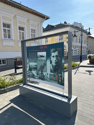 This is a medium shot of an outdoor informational display in the city of Nikul'skoe (Neverkovskii S/O), Russia. The display is housed in a grey metal frame and features a large banner with text and images. The banner appears to be a memorial or historical exhibit, with a portrait of a man in military uniform, along with other photographs of military scenes and medals. The text on the banner is in Russian, with the title "- НАШИ ГЕРОИ -" (Our Heroes) prominently displayed.
The display is situated on a paved walkway made of grey interlocking bricks. To the left of the display, there is a dark green park bench. Behind the display and bench, there is a building with a pale yellow facade and white decorative elements around the windows and roofline. Further in the background, other buildings with similar architectural styles can be seen, one of which is painted yellow with white ornamentation.
The scene is illuminated by bright sunlight, casting strong shadows on the ground. Green bushes are planted along the edge of the walkway. The sky is clear and blue. The overall impression is of a well-maintained public space, likely a town square or a pedestrian street.