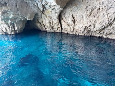 The image depicts a view from a boat entering a sea cave on Antipaxos, Greece. The water is a vibrant, clear turquoise, and the cave opening is visible on the left, revealing a darker interior. The cave walls are composed of rough, textured rock, predominantly light gray and beige, with some areas appearing eroded or layered. Sunlight illuminates the rock faces, creating highlights and shadows, and also reflects off the water's surface, causing ripples and shimmering patterns. The overall scene conveys a sense of natural beauty and exploration. The time of day appears to be daytime, with bright natural lighting suggesting fair weather. There are no people or text visible in the image. The dark shape in the water is a reflection or shadow, possibly of the boat from which the photo was taken.