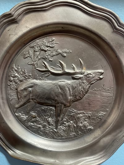This image shows a close-up of a decorative metal plate with a bas-relief scene of a stag. The stag is depicted in profile, facing right, with its mouth open as if roaring or calling. It has large antlers and is standing in a wooded area with a tree behind it and water or a field in front. The plate has a scalloped edge. The object appears to be made of pewter or a similar metal. The background is a light blue surface. No specific location in Schwäbisch Hall, Germany, can be identified from this image.