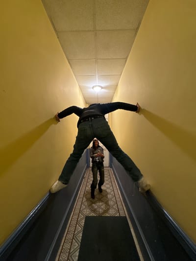 A narrow, pale yellow-walled hallway in Roanne, France, is depicted from a low, wide-angle perspective. In the foreground, a person is seen from behind, bracing themselves horizontally between the two walls, with their arms and legs spread wide in an 'X' shape. This individual wears dark blue jeans, a dark jacket, and light-colored footwear. Directly ahead, framed by the foreground person's legs, a second individual stands facing the camera. This person, identifiable as a female with dark hair and a smile, is dressed in a dark jacket, dark pants, and dark boots. She holds a white smartphone horizontally with both hands. The hallway floor features a patterned tile design in beige and brown tones, bordered by a dark blue skirting board. A dark rectangular mat lies on the tiled floor in the foreground. The ceiling is a suspended acoustic tile grid, illuminated by a single, central, circular flush-mount light fixture.