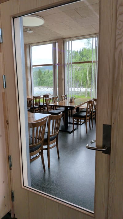 This is a photograph taken through an open doorway, looking into a restaurant or dining area. The room features wooden tables and chairs with dark upholstery, arranged in a setting that suggests a casual dining establishment. Large windows line one wall, offering a view of a scenic landscape with a river, lush green trees, and hills under an overcast sky. The lighting appears to be natural, coming from the windows and possibly overhead fixtures, indicating it is daytime. The overall atmosphere is serene and inviting, with a sense of natural beauty. The doorframe and the interior of the room are constructed with light-colored wood, contributing to a warm and rustic aesthetic. The floor is a dark, speckled material. No people are visible in the image, suggesting the restaurant is either closed or not yet open for service. The location is identified as Trofors, Norway, which aligns with the natural, possibly Scandinavian, landscape depicted through the windows.