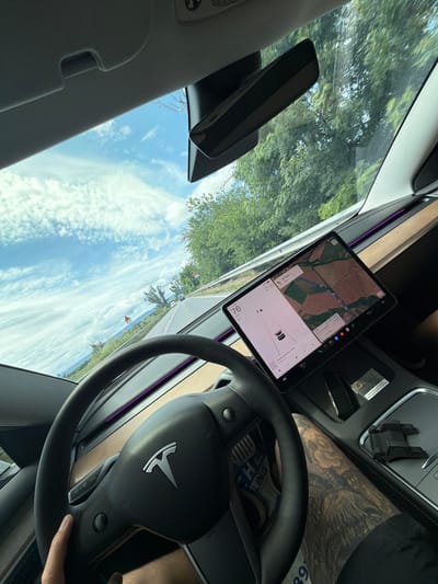 The image shows the interior of a Tesla car, taken from the driver's perspective. The driver's left hand is on the steering wheel. The car is in motion on a road, indicated by the landscape visible through the windshield, which includes trees, a road, and a cloudy sky with blue patches. The car's central touchscreen display is visible, showing navigation information with a route highlighted on a map. The dashboard has wood-grain accents and ambient purple lighting. On the center console, there's a phone mount and what appears to be a small silver and black object. The driver's legs are visible, and they have intricate tattoos on their right leg. The scene implies the car is traveling on a rural or semi-rural road. The image is dated to August 2023. There is no specific indication of Ivanski, Bulgaria within the image.