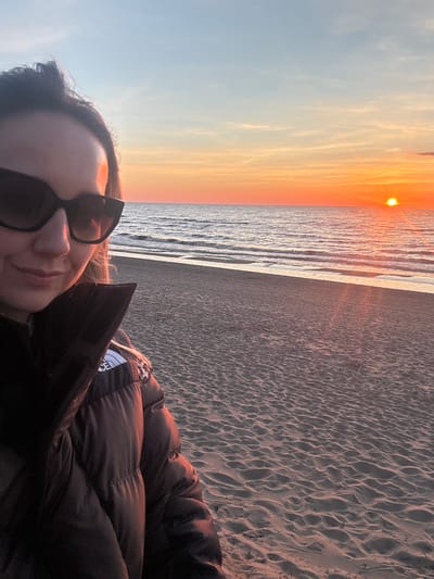A person wearing a black puffer jacket and sunglasses is seen in a selfie taken at a beach at sunset. The beach is sandy and the sea reflects the sunset colors. The sky is transitioning from blue to orange near the horizon. The location is Garupe, Latvia.