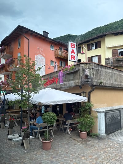 In the city of Fai della Paganella, Italy, an outdoor seating area of a bar and B&B is depicted. The establishment features an orange building on the left with the inscription "B&B Casa Agostini" and a mural. To its right is a yellow building. A prominent red "BAR" sign hangs vertically. The area is furnished with wooden tables and chairs, some covered by white umbrellas. Several patrons are seated at the tables. A man in a blue shirt and jeans sits at one table, while another man with a shaved head is seen from behind at a different table. A woman with dark hair and glasses sits with the man in the blue shirt. A tree stands near the seating area, and potted plants are strategically placed throughout the scene. The ground is paved with cobblestones. The sky is overcast, and a forested mountain slope is visible in the background.