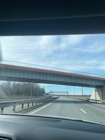 The image shows a view from inside a vehicle traveling on a highway. The road has multiple lanes separated by painted lines and a concrete barrier. An overpass is visible above the highway. The environment includes trees lining the side of the road and a partially cloudy sky. A semi-trailer truck can be seen on the highway. The location is Kliuchiki, Russia.
