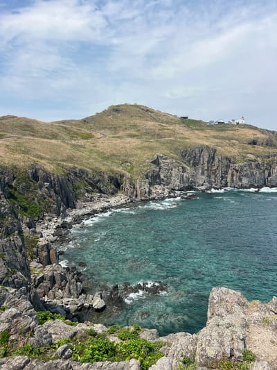 The image presents a view of a coastal cliff landscape. The foreground features a rocky outcrop with some green vegetation. The water along the shore is turquoise and relatively clear, with waves visible. A hillside covered in grass and some exposed rock faces rises from the shoreline, sloping upwards towards the top left of the frame. Buildings can be seen at the top of the hillside. The sky is overcast. The scene is of Mayak Byusse, Russia.