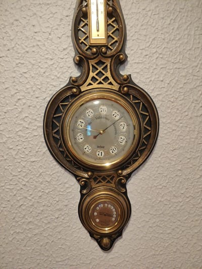 The image displays a decorative, ornate wall-mounted barometer and thermometer. The main barometer dial has Roman numerals and has a pointer indicating "Kaunista." The barometer is made of brass and dark wood with gold accents. Above the main barometer, there is a thermometer with markings from 0 to 10. Below the main barometer, there is a smaller dial, likely for humidity or another atmospheric measurement, with a pointer and markings from 0 to 100. The entire unit is mounted on a textured white wall. The objects are stationary, and no people are present. The overall impression is of an antique instrument.
This object is located in the Kymi Museum in Kotka, Finland, as indicated by the text "Kymeen Museossa" visible on the barometer dial, which translates to "In the Kymi Museum."