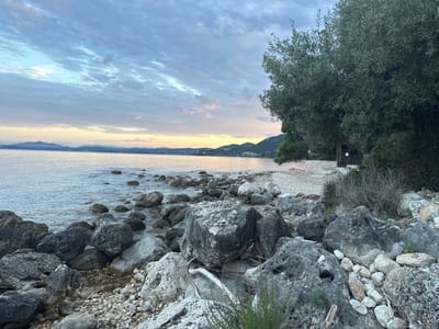 A wide-angle, eye-level shot captures a rocky beach scene at dusk in Kassopaia Municipal Unit, Greece. The sky above is filled with a mosaic of grey and blue clouds, tinged with pale pink and yellow hues from the setting sun near the horizon. A distant coastline with low hills is visible across a calm body of water. The foreground is dominated by a collection of large, grey and white boulders and smaller pebbles. A sparse, olive tree with dense foliage arches over the scene from the right side, casting shadows. In the middle ground, a small, rustic wooden structure with a metal roof is nestled amongst vegetation and stone walls, suggesting a secluded spot. The overall atmosphere is serene and natural, with no people or animals present.