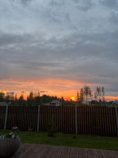 The image depicts a rural or suburban scene at dusk, possibly in Gubakha, Russia. The sky is dominated by clouds, with a vibrant sunset casting orange and pink hues across the horizon. Silhouetted trees and buildings are visible against the colorful sky, including what appears to be a church spire. In the foreground, a dark wooden fence separates a grassy yard from the distant landscape. A woven planter with white flowers sits to the left of the fence, and a wooden deck is visible at the bottom of the frame. The overall impression is one of a peaceful evening in a quiet residential area.