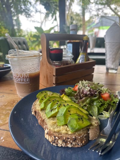 On a wooden table in Baan Kai, Thailand, a partially eaten plate of avocado toast with a side salad sits next to a glass of iced coffee. The toast is topped with sliced avocado and black sesame seeds, and the salad contains lettuce and chopped tomatoes. A fork is placed on the plate, ready to eat. A wooden stand holds various bottles and containers. The foreground features a cup of coffee with condensation and a plastic straw. The background is blurred, showing greenery and structures, indicating an outdoor setting, likely a cafe or restaurant.