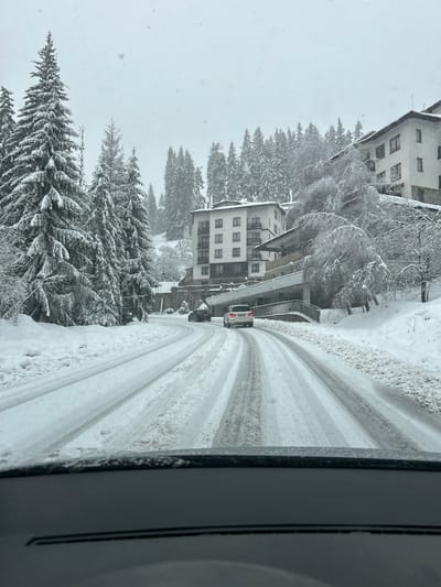 The image shows a snowy scene, possibly in Pamporovo, Bulgaria, given the presence of buildings consistent with alpine architecture and heavy snowfall. A road curves through the scene, marked with tire tracks indicating recent vehicular traffic. Snow-covered pine trees line the side of the road. Buildings with multiple floors and windows are visible among the trees. The weather appears to be actively snowing. A vehicle is visible on the road. The image seems to be taken from inside a building or vehicle.