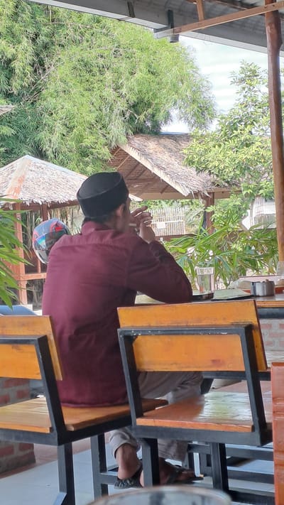 A man wearing a maroon long-sleeved shirt and a black peci hat sits at a table outside under a wooden overhang. He is facing away from the camera and appears to be smoking a cigarette, holding it in his right hand near his face. He is wearing light-colored pants and dark sandals. In front of him, on the table, are a glass of water, a notebook, and an ashtray. Behind the man, a motorcycle helmet is visible. The background consists of lush green foliage, including bamboo, and thatched-roof structures. The overall setting suggests an outdoor seating area, possibly a cafe or restaurant, in Julikeude Dua, Indonesia.