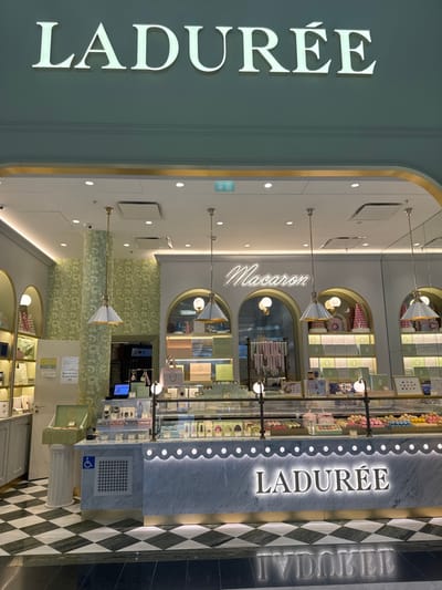 The image depicts the interior and immediate entrance of a Ladurée boutique, likely located within Paris Charles de Gaulle Airport (CDG) in Roissy-en-France, France.
The scene presents a brightly illuminated, unoccupied retail space specializing in macarons and confectionery. Above the storefront, a prominent, backlit sign reads "LADURÉE" in white capital letters. Inside, a white neon sign spells "Macaron" in script.
The interior features a light green and white color palette with gold trim accents. The rear wall transitions from light green patterned wallpaper on the left to white panels with multiple arched display niches. These niches showcase various boxed goods, macaron pyramids, and decorative confectionery. Several white and gold-toned pendant lights hang from the white ceiling, which also contains recessed lighting and air vents.
A long, white marble-clad counter with a scalloped, backlit LED trim occupies the lower portion of the storefront. A backlit "LADURÉE" sign in white letters is centered on the front of this counter. Glass display cases on top of the counter contain an assortment of colorful macarons, pastries, and other packaged confections. On the left side of the counter, a blue international accessibility symbol (wheelchair icon) is affixed to a grey gridded panel. To the far left, an open shelving unit displays additional packaged goods.
The floor in the immediate foreground features a black and white checkered tile pattern, transitioning to a darker, solid-colored floor closer to the viewer. No individuals are present in the scene.