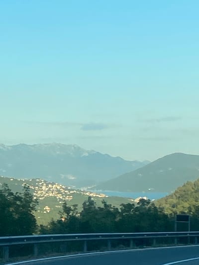 The image captures a scenic view from a high vantage point, likely along a road, looking out over a valley and distant mountains. In the foreground, a metal guardrail runs horizontally across the lower part of the frame, separating a road from the view beyond. Lush green trees and foliage line the slope below the guardrail, obscuring the immediate foreground.

Further in the distance, a town or village is nestled on the hillside, characterized by numerous small, brightly lit structures that stand out against the green landscape. This settlement appears to extend down towards a body of water, possibly a bay or inlet, which is visible in the mid-ground. The water reflects the light and appears calm.

Beyond the water, a range of mountains stretches across the horizon. These mountains are hazy and muted in color, with some peaks appearing snow-capped or covered in light-colored clouds. The sky above is a clear, gradient blue, transitioning from a lighter shade at the horizon to a deeper blue towards the top of the frame.

The scene suggests a picturesque location, possibly a Mediterranean coastal region given the terrain and vegetation. The presence of the road and guardrail indicates a developed area accessible by vehicle, offering panoramic views to travelers. While the specific location "Palje Brdo, Croatia" is mentioned, the image itself does not contain explicit signage or landmarks that definitively confirm this location. However, the visual elements are consistent with the geographical features found in such coastal mountainous regions of Croatia.