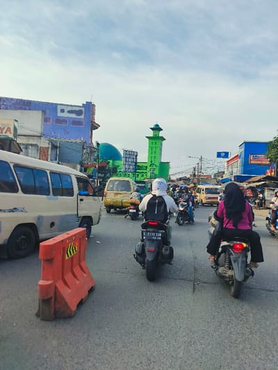 The scene is a busy street in Kecamatan Dayeuhkolot, Indonesia, located at coordinates -6.99 latitude and 107.627 longitude. A large white van is parked on the left side of the road with a red and yellow barrier in front of it. There are two motorcycles on the right side of the road, one is a black motorcycle being driven by a man wearing a black helmet and jacket, and the other is a black motorcycle being driven by a woman wearing a pink hijab. There are several other vehicles, including cars and motorcycles, visible in the background of the scene. The scene is framed by buildings on both sides of the street. In the center of the image, a mosque with a green dome and minaret can be seen. The sky is partly cloudy with a blue and white hue. The ground is paved with asphalt.