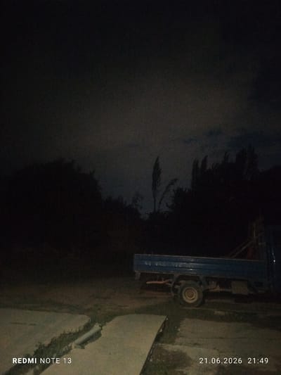 In the city of Асан-Чек, Kyrgyzstan, at 21:49 on June 21, 2026, a dimly lit outdoor scene captures a parked blue truck. The truck is positioned on a dirt and grassy area, with a concrete walkway or slab in the foreground. The background consists of dark silhouettes of trees against a cloudy night sky. The truck appears to be a utility or light cargo vehicle, with its bed empty. The overall atmosphere is dark and quiet, suggesting it is nighttime in a rural or semi-rural setting. The image contains embedded text indicating "REDMI NOTE 13" and the date/time.