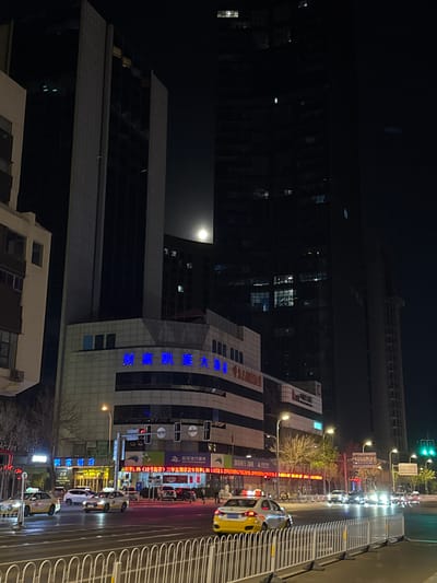 This nighttime image captures a bustling urban street scene in Shenhe District, China. The setting features multiple tall buildings, including dark glass-faced skyscrapers with scattered illuminated windows, and a prominent lighter-colored building adorned with blue illuminated Chinese characters. The moon is visible high in the dark sky, casting a subtle glow.

The street is busy with traffic, including several cars and distinctive yellow and white taxis. Concrete barriers and white metal fences line the side of the road, separating the pavement or a pedestrian area from the vehicular traffic. Streetlights cast pools of light, and a green traffic light is visible, indicating active traffic flow.

Visible text on the buildings and street-side signs provides specific location and commercial information. The most prominent text, in blue illuminated characters, reads "财富凯莱大酒店" (Cáifù Kǎilái Dà Jiǔdiàn), which translates to "Fortune Kai Lai Grand Hotel" or "Wealth Kai Lai Hotel." Lower down, another sign shows "Archer V Hotel" and "凯莱酒店" (Kǎilái Jiǔdiàn), likely indicating the same hotel or a related establishment.

Other signs display financial information, including "自助银行服务" (Self-service Banking Services) and an advertisement for "三年定期存款年利率1.9%(10万起存)" (Three-year fixed deposit annual interest rate 1.9% (starting from 100,000)), suggesting the presence of a bank or financial institution. The numbers "1905" and "78" are also visible on signs, possibly indicating addresses or other identifiers.

The overall impression is of a vibrant, modern city environment at night, characterized by artificial lighting, architectural contrasts, and active urban life.