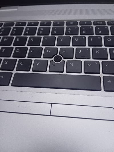 The image displays a close-up, angled view of a laptop keyboard and a portion of its touchpad, rotated 90 degrees clockwise. The scene is likely set indoors in Blantyre, Malawi.

The central subject is a dark grey/black QWERTY keyboard with white lettering. Keys visible include the full alphabet from 'Q' through 'M', along with the number row (1 through 0) and their corresponding symbols such as '!', '@', '#', '$', '%', '^', '&', '*', '(', ')'. Key labels like 'Alt' are also clearly seen. A prominent feature is a circular, textured black trackpoint (pointing stick) positioned between the 'G', 'H', and 'B' keys.

To the right, a silver-colored touchpad is visible, featuring a subtle horizontal demarcation for its click zones. The laptop's casing, also silver/light grey, frames the keyboard and touchpad, showing a clean surface with minimal dust or wear. No specific activity is occurring, but the image focuses on the input devices of a functional laptop.