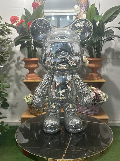 In the city of Novo-Glagolevo, Russia, a life-sized, silver, disco ball-covered bear statue stands prominently on a black marble-topped table with a gold trim. The statue is positioned in front of a white, paneled wall and a mirror. Behind the statue, two large potted plants with dark green leaves and red flowers are visible, flanking a smaller potted plant on a wooden shelf. To the right of the statue, a light purple woven basket filled with assorted flowers is placed on another wooden shelf. The floor in front of the table is covered with green artificial turf.