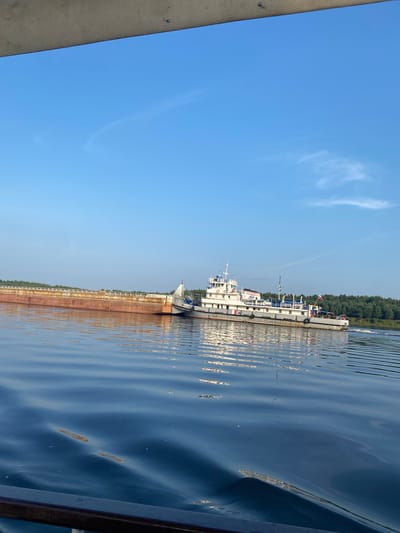 A towboat is pushing a barge along a river, with a dense forest on the far bank under a clear blue sky. The towboat, painted white with red and blue accents, has a superstructure with multiple decks and windows, and a Russian flag is visible at its stern. The barge, a long, flat-bottomed vessel with rust-colored sides, appears to be carrying cargo. The water is calm with gentle ripples, reflecting the blue of the sky and the white of the boats. The scene suggests a sunny day with good visibility. The location is specified as the city of Поварёнки, Russia.